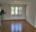 Empty room with wooden flooring, large windows, radiator, and a small table with a few items in the corner.