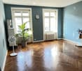 Empty room with wooden flooring, blue walls, large windows, a fan, a potted plant, and a small table with a vase of flowers.