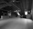 An empty, spacious room with exposed wooden beams and a high, slanted ceiling. The room features large windows allowing natural light to enter, and a small kitchen area is visible in the background.