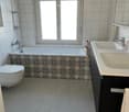 Modern bathroom with a bathtub, toilet, double sink, and a large window. The bathtub features a decorative tile border, and the sink has a modern faucet. A towel is visible on the cabinet.