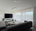 Modern living room with a grey sectional sofa, coffee table, television, fan, and small round table near large windows with sheer curtains.