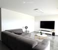 Modern living room with a grey sectional sofa, a white coffee table, a flat-screen TV mounted on the wall, and a small side table with a fan.