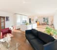 Bright, open-plan living and dining area with wooden flooring, white kitchen island, dark blue sofa, red armchair, glass coffee table, and dining area with chairs and table.