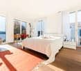 Bright bedroom with large windows offering a scenic view, featuring a bed with white bedding, a red accent chair, a wooden desk with a chair, and a nightstand with a lamp.