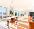 Bright, open-plan living and dining area with wooden flooring, large windows offering a scenic view, a dining table with chairs, a vase of flowers, a kitchen area with a counter, sink, refrigerator, and potted plants.