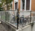 A white metal fence encloses a small outdoor area with several bicycles parked inside. The fence is attached to a concrete platform, and the background shows a multi-story brick building with windows and shutters.