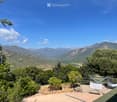 A scenic outdoor area with a panoramic mountain view, surrounded by lush greenery and trees, featuring a patio with lounge chairs and a small table.