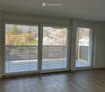 Empty room with wooden flooring and large windows with blinds, offering a view of the outdoors.