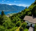 A small white house with a tiled roof, surrounded by lush green trees and shrubs, overlooking a serene lake and distant mountains.