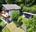 A two-story house with a gray roof and white walls, featuring a wooden balcony and outdoor seating area surrounded by greenery.