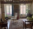 Cozy living room with wooden floor, exposed wooden ceiling beams, large windows, green sofa, white armchair, wooden table, colorful rug, and various plants.