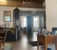 A cozy, well-lit room with wooden beams on the ceiling, a large window, and a mix of modern and traditional furniture including a chair, a round stool, a wooden table, a piano, and a ball on the floor.