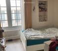 Cozy bedroom with a single bed, nightstand, radiator, built-in shelf, and large windows with curtains.