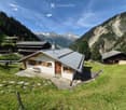 A small white wooden house with a grey metal roof, wooden shutters, and a wooden door, situated on a grassy slope with a stone pathway leading to the entrance. The surrounding area features other wooden houses, lush greenery, and distant mountain peaks.