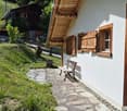A white wooden house with a slanted roof and wooden shutters, featuring a stone pathway leading to a wooden bench, surrounded by greenery and a wooden fence.