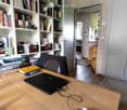 A well-organized home office with a wooden desk, laptop, chair, bookshelf filled with books, and a small plant in the background.