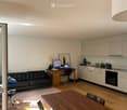 A modern, open-concept living and dining area with a black leather sofa, wooden desk with laptop and monitor, dining table with chairs, and a kitchen area with cabinets and appliances.