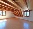 Empty attic room with wooden beams, wooden flooring, and two windows offering a view of the roof and surrounding area.