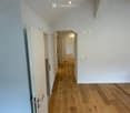 Empty hallway with wooden flooring, white walls, and recessed lighting. Features an open doorway leading to another room.