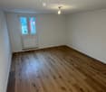 Empty room with light-colored wooden flooring, white walls, and a single window with a radiator below it.