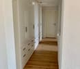 Clean, minimalist hallway with light wooden flooring, white walls, built-in white storage units with mirrored doors and drawers, and a closed door at the end.