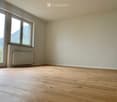 Empty room with light wooden flooring, white walls, large window with view, radiator, and power outlets.