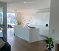 Modern open-plan living and dining area with a kitchen island, white cabinets, a sofa, a coffee table, and a dining table with chairs. The space features wooden flooring, large windows with a city view, and decorative plants.