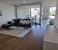 Modern living room with wooden flooring, large windows, a black sofa with white and red cushions, a glass coffee table, side tables with plants, and a mirror on the wall.