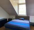 Cozy attic bedroom with wooden slanted ceiling, large window, bed with colorful bedding, nightstand, lamp, and small chair.