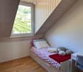 Cozy attic bedroom with a single bed, colorful bedding, pillows, and a small desk with a chair. Large window offers a view of a vineyard.