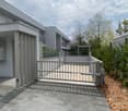 Modern residential building entrance with a gated driveway, grey stone tiles, and surrounding greenery.