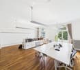 Bright, modern office space with wooden flooring, large white table, several chairs, cabinet with books, laptop, and plants near the window.