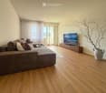 Modern living room with a sectional sofa, coffee table, flat-screen TV on a wooden stand, floor lamp, and decorative vase with dried branches.