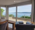 Bright, modern living space with large windows offering a scenic view of a lake. Features a wooden dining table with chairs, a leather sofa, and a small bench near the sliding glass doors leading to a balcony.