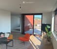 Modern living room with wooden flooring, large sliding glass doors leading to a balcony, a black sofa with decorative pillows, a small black table with a red lamp, an orange armchair, and a large potted plant near the window.
