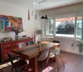 Bright dining area with a long wooden table surrounded by modern chairs, a sideboard with decorative items, a lamp, and a large painting on the wall. Large windows allow natural light to fill the space.