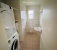 Bathroom with a toilet, sink, washing machine, dryer, and shelves. The room has tiled walls and floor, and a small window providing natural light.