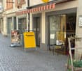 A narrow cobblestone street lined with historic buildings, featuring a storefront with a striped awning, a yellow signboard, and a small outdoor seating area with a table and chairs.