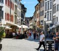 A bustling cobblestone street lined with historic buildings, outdoor seating areas, and pedestrians. The street is adorned with street lamps, signage, and a sign advertising a coffee bar.