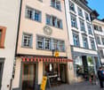 A multi-story building with a light-colored facade, multiple windows, and a striped awning over the entrance. The ground floor features a shop with visible tables and chairs inside, and a yellow signboard outside.