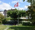 Well-maintained garden with lawn, flower beds, shrubs, trees, patio seating area, and Swiss flag on a pole.