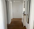Clean, minimalist hallway with light wooden flooring, white walls, and a small white cabinet with a mirror and decorative items on top.