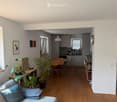 Open-plan living and dining area with a sofa, coffee table, dining table, chairs, and a kitchen area with cabinets, appliances, and a window. The space features wooden flooring, large windows, and several potted plants.
