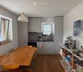 Modern kitchen with light grey cabinets, wooden dining table, and various appliances including a coffee maker and oven. The space features large windows with blinds and a pendant light hanging from the ceiling.