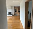 Spacious hallway with wooden flooring, white walls, and a fireplace on the left side. A kitchen area is visible in the background with wooden cabinets and stainless steel appliances.