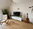 Modern living room with wooden flooring, white walls, and slanted ceiling. Features a white armchair, a guitar, a television on a white cabinet, a vase with flowers, and speakers.