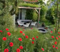 A lush garden with red poppies, a wooden pergola, white chairs, and a table, surrounded by greenery and trees.