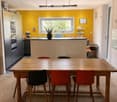 Bright kitchen with a yellow accent wall, wooden dining table, and modern appliances. Features include a bar counter, stools, and a potted plant on the counter.