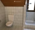 Small bathroom with a toilet, bathtub, and a window with a showerhead above it. The walls and floor are tiled in light beige.