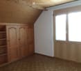 Empty attic room with wooden walls, slanted ceiling, large window, and built-in wooden cabinet with shelves.